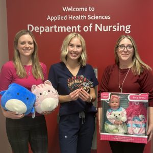 Three women stand in a row in front of a red wall that reads "Welcome to Applied Health Sciences Department of Nursing." The first woman holds two large stuffies. The second women fans out several gift cards. The third woman holds a baby doll toy.