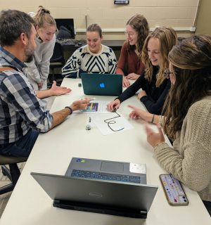 A group of female Brock students sit around a table with a man in a blue patterned shirt, looking at a laptop while. The students are all smiling looking at the educational materials.