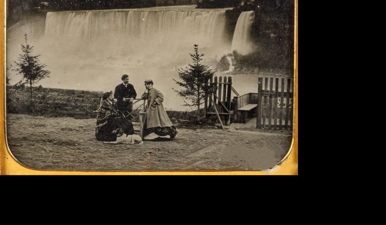 A black and white photo of two women, a man and a dog dressed in clothing from the mid 1800s standing posing for a photo with a view of Niagara Falls in the background.