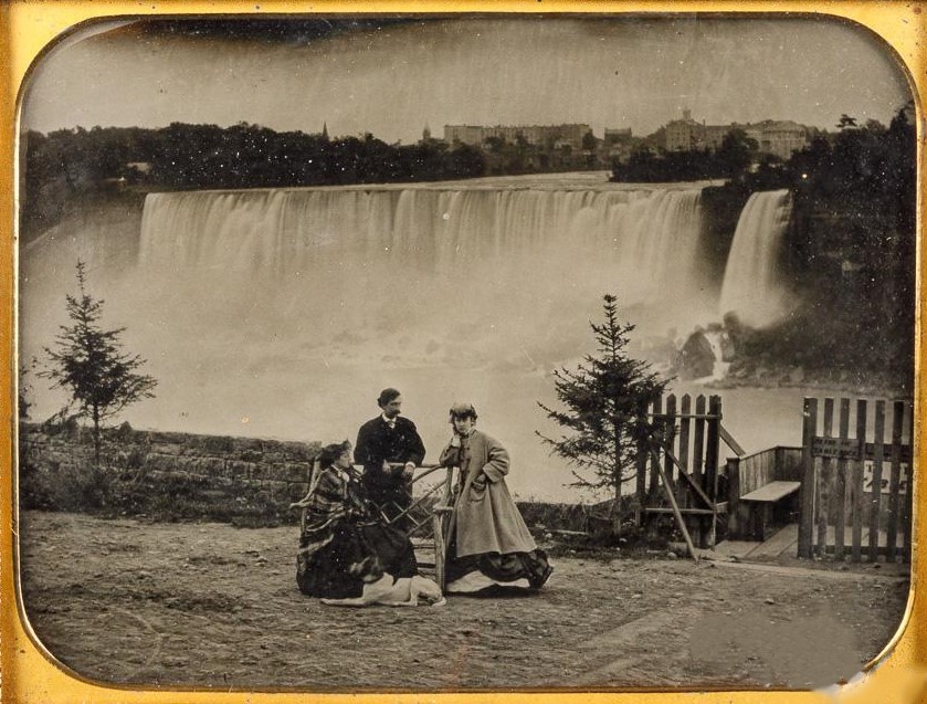 A black and white photo of two women, a man and a dog dressed in clothing from the mid 1800s standing posing for a photo with a view of Niagara Falls in the background.