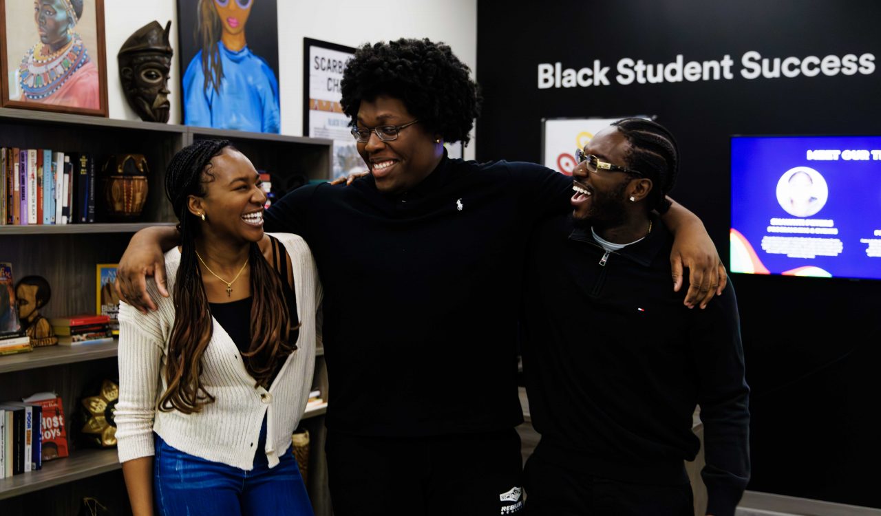 Three students stand with their arms around each other in front of a wall sign that says ”Black Student Success.”