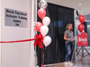 A woman speaks at a podium with a “Brock University” logo on it. A sign with “Brock Functional Inclusive Training Centre” can be seen on a wall near her.