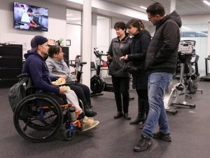 A man and woman in a wheelchair speak to three people in a spacious exercise facility.