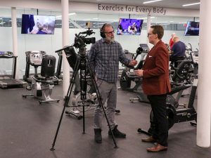 A man with a video camera and microphone interviews another man in a spacious exercise facility.