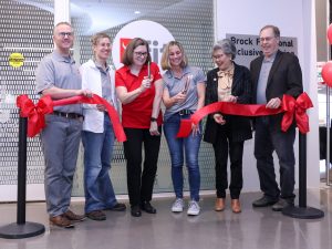 Six people stand behind a long, horizontal red ribbon in front of a building with signage that reads “Bfit, Brock Functional Inclusive Training Centre.” Two people in the centre of the group use scissors to cut the ribbon.