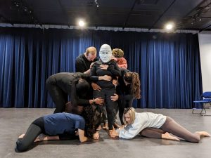 Brock University Dramatic Arts students rehearse in a theatre space with a black stage and blue curtain in the background. They stand in a group surrounding an actor with a white mask over her face.