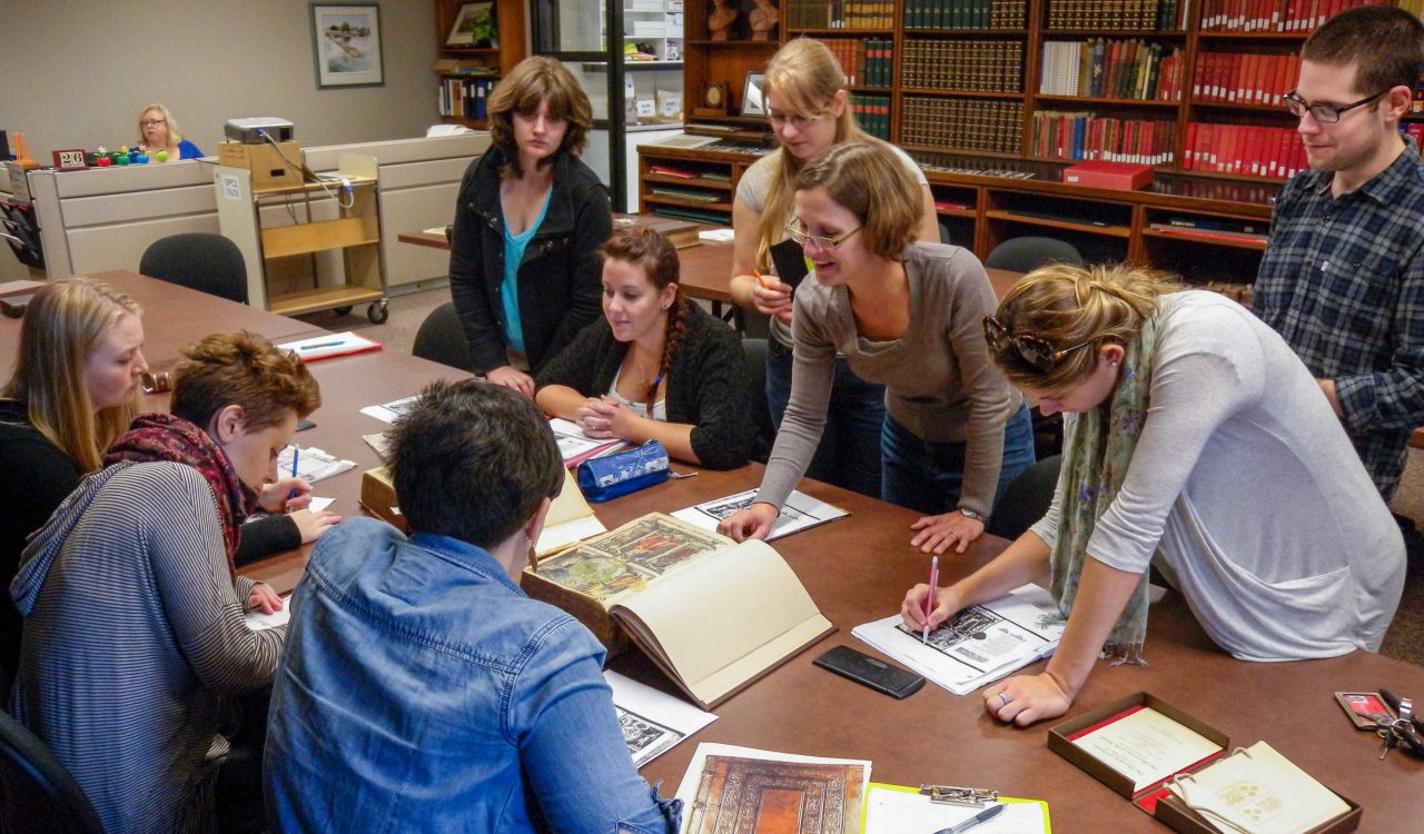 A group of Brock University students huddle around a table in Brock’s Archives and Special Collections looking at historical texts. Professor Leah Knight leans over a book smiling.
