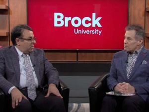 Two men sit in chairs in front of a television screen showing the Brock University logo.