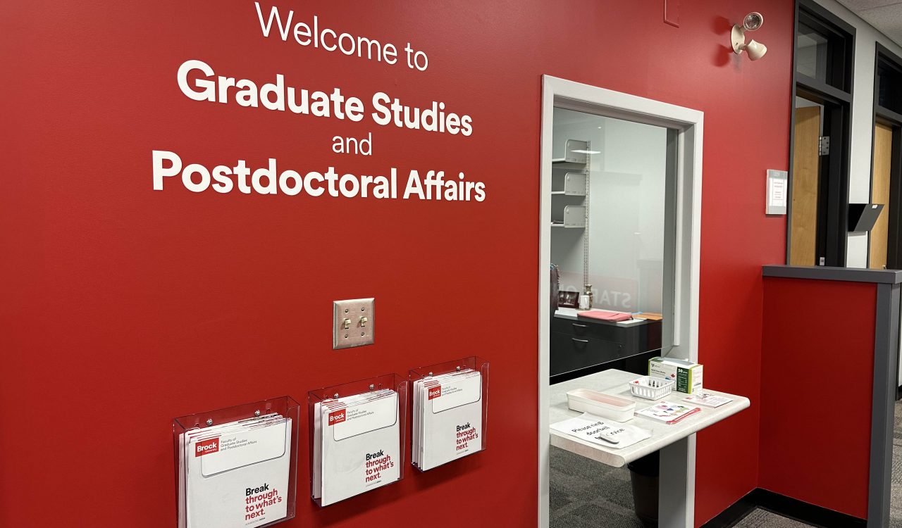 The words "welcome to graduate studies and postdoctoral affairs" painted in white on a red wall beside an office door. Containers full of brochures are attached to the wall.