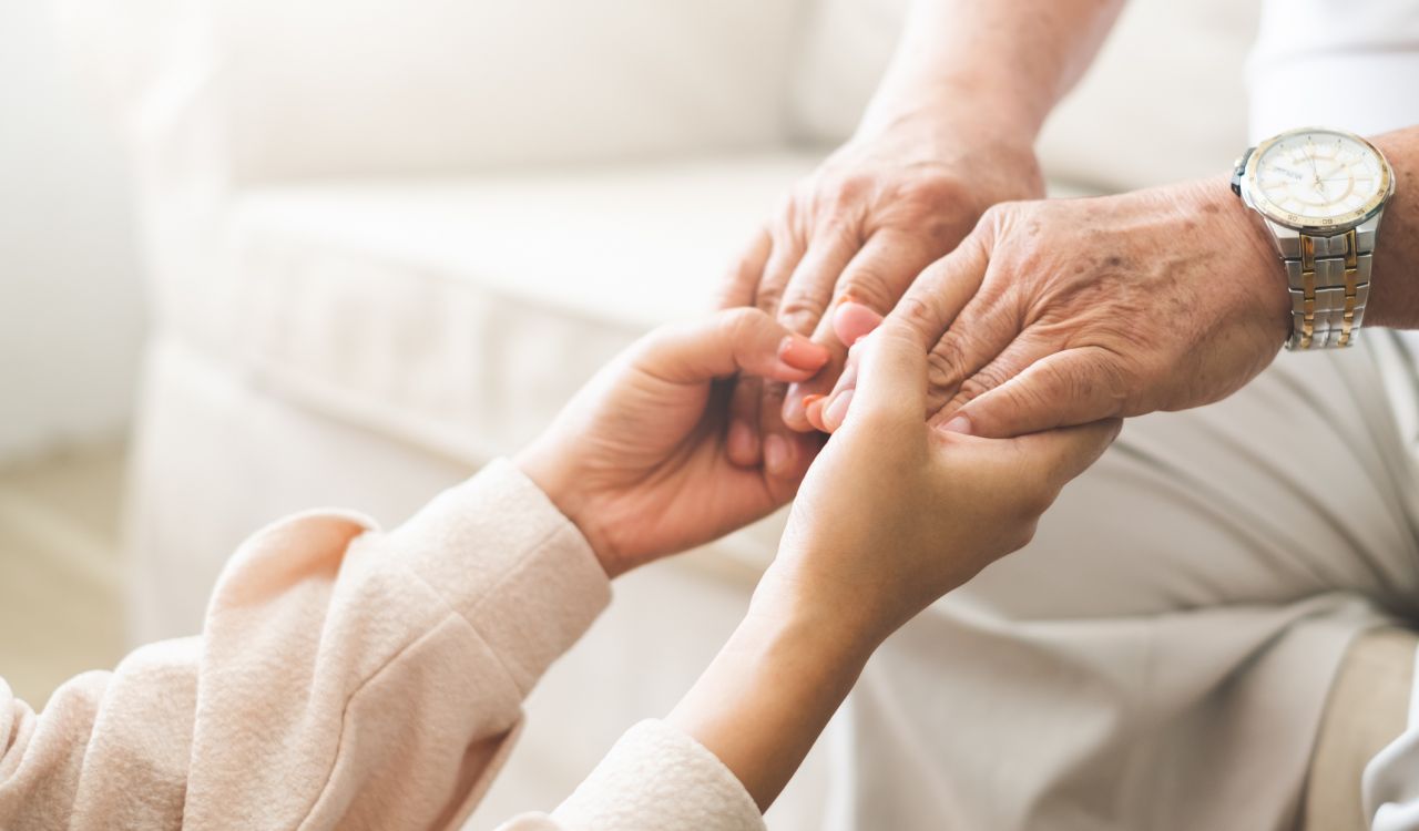 Close-up of a young person holding an elderly person's hand in a sunny living room.