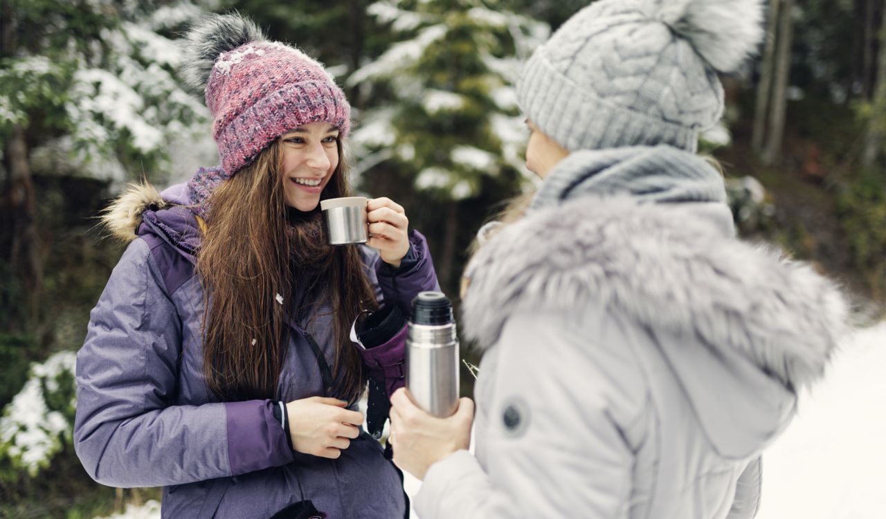 Two people walk outside in the snow on a winter day. They are dressed in winter gear and drinking from thermos flasks.