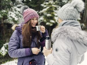 Two people walk outside in the snow on a winter day. They are dressed in winter gear and drinking from thermos flasks.