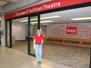 A woman wearing a red shirt stands below a sign that says Sean O’Sullivan Theatre