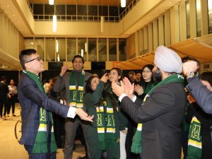 Several Alberta School of Business MBA students wearing faculty-branded scarves raise their arms and clap while one of their team members records the team cheering on a phone.