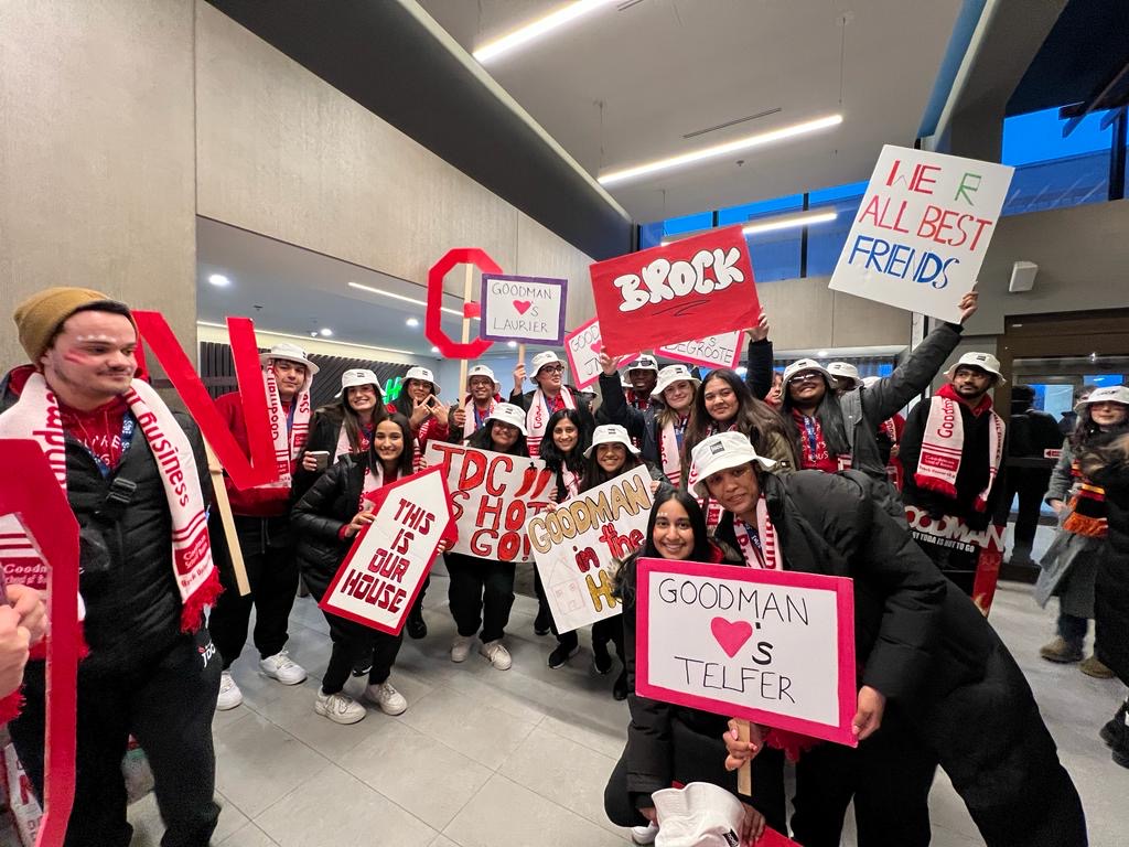University students dressed in white bucket hats and red and white scarves stand in an entryway holding spirit signs.