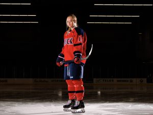 A female hockey player in a red jersey stands fully equipped on the ice.