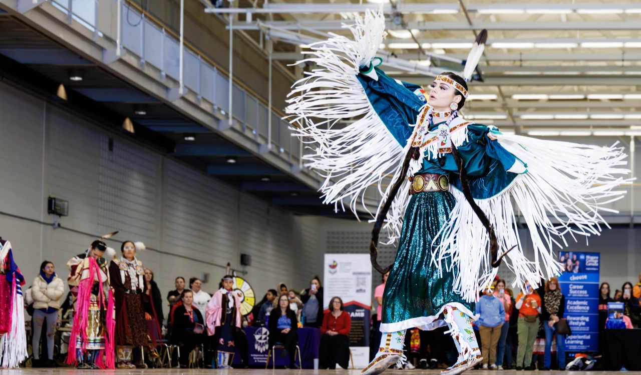 A woman in traditional Indigenous regalia dances in front of a crowd in a gymnasium.