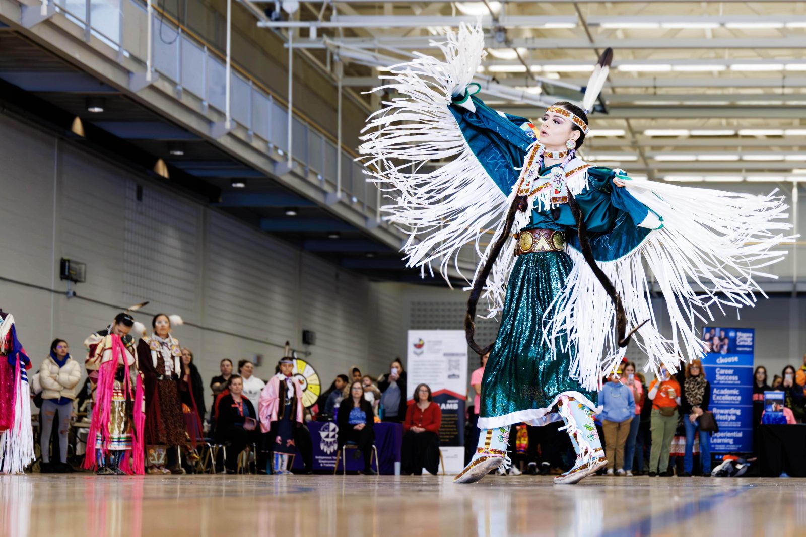A woman in traditional Indigenous regalia dances in front of a crowd in a gymnasium.