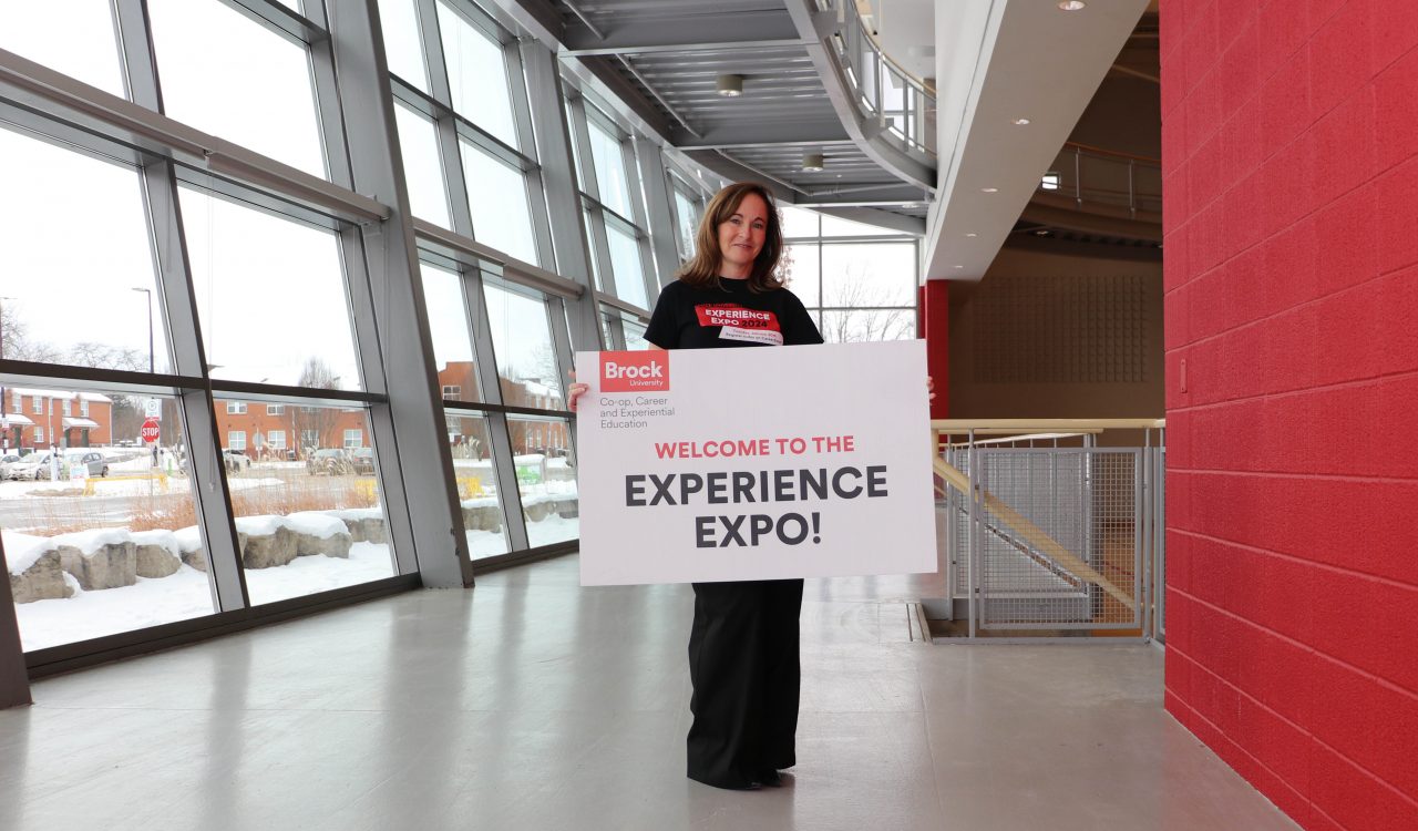 A woman stands next to a university gymnasium holding a sign that says Welcome To The ‘Experience Expo’
