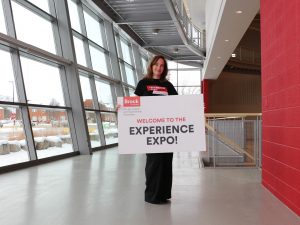 A woman stands next to a university gymnasium holding a sign that says Welcome To The ‘Experience Expo’