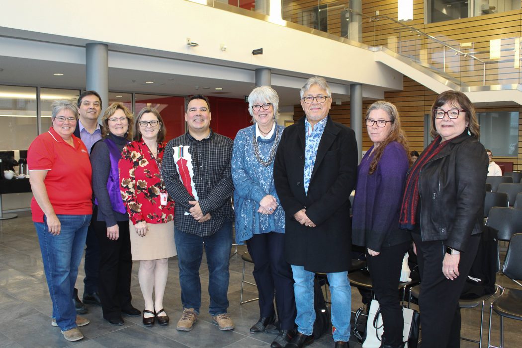 A group of faculty members and staff from Brock University alongside Indigenous Scholar Niigaan Sinclair stand arm to arm in Brock’s Atrium in the Goodman School of Business. They smile warmly at the camera. 