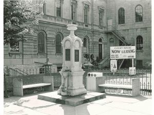 A fountain sits at the corner of a city street. There are two benches on each side. In the background is a building and a large sign that says, courthouse now leasing office retail space.