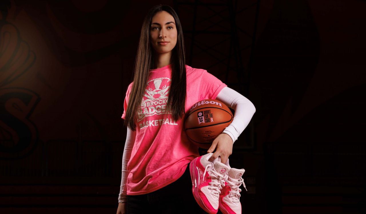 Olivia Fiorucci, a Brock University student-athlete, stands holding a basketball and a pink pair of shoes with breast cancer awareness messaging.