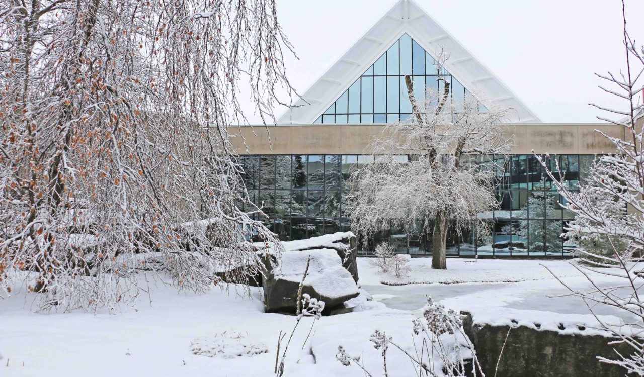 A large building with triangular glass windows sits behind a snow-covered pond.