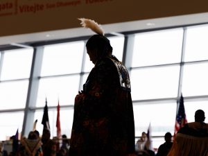 A person in traditional Indigenous regalia is silhouetted against a wall of windows at a powwow.
