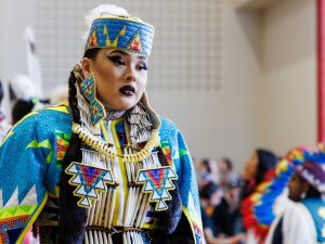 A woman wears traditional Indigenous regalia at a powwow. A crowd is visible in the background.