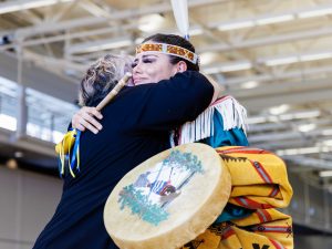 Two women hug in a gymnasium. One is wearing traditional Indigenous regalia.