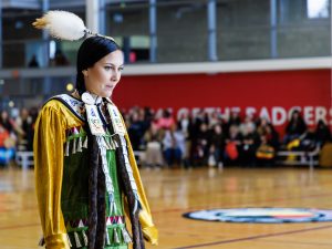 A woman in traditional Indigenous regalia stands in a gymnasium.