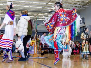 People in traditional Indigenous regalia dance in a gymnasium as part of a Pow Wow.