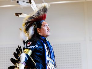 A man in traditional Indigenous regalia stands in a gymnasium.