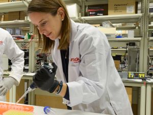 Associate Professor of Health Sciences Rebecca MacPherson looks at a tray of test tubes as she leans over a counter in the lab, holding an instrument in her gloved hand.