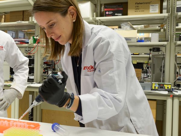 Associate Professor of Health Sciences Rebecca MacPherson looks at a tray of test tubes as she leans over a counter in the lab, holding an instrument in her gloved hand.