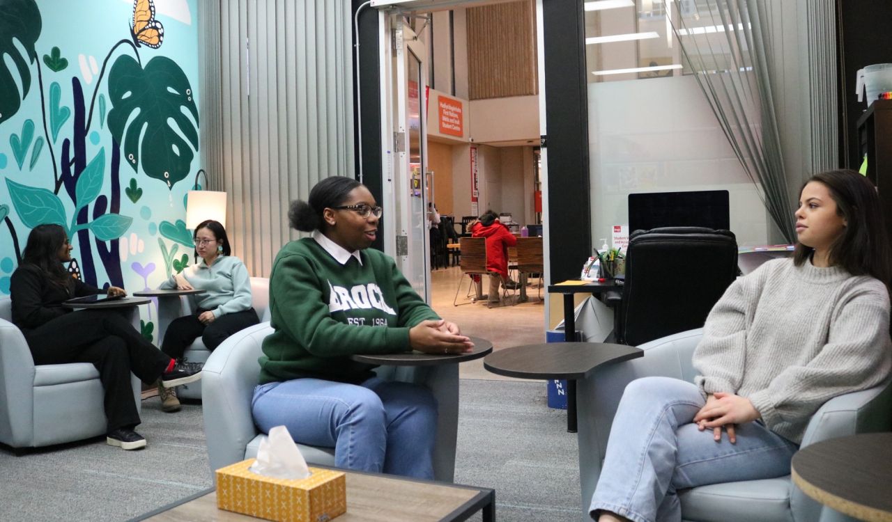 Two female students sit in chairs having a conversation. In the background two female students sit on either side of a table talking to each other.