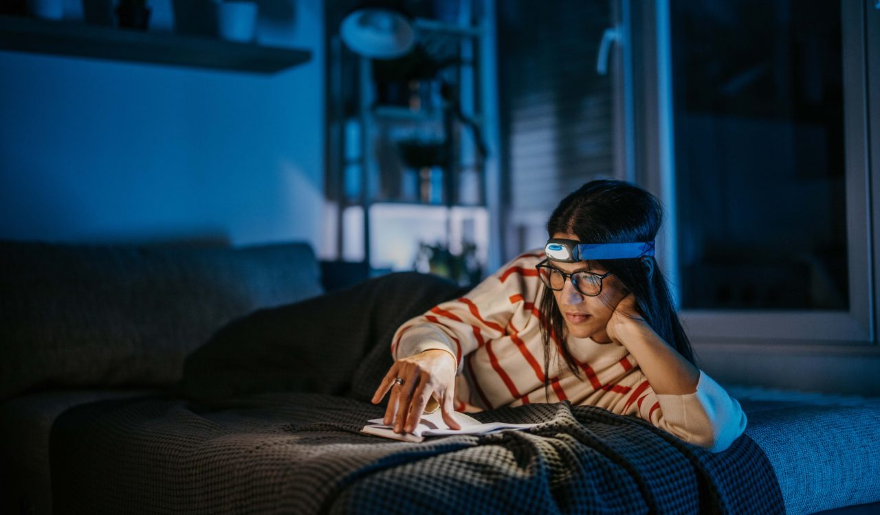 A woman reads on her bed in the dark, while using a head lamp to save electricity.