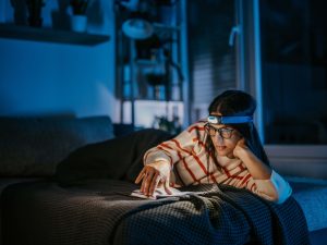 A woman reads on her bed in the dark, while using a head lamp to save electricity.