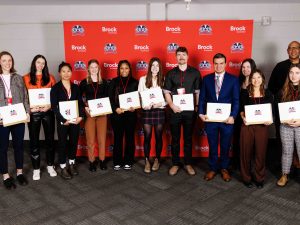 A group of student-athletes stand holding their academic awards in front of a Brock University-branded backdrop.