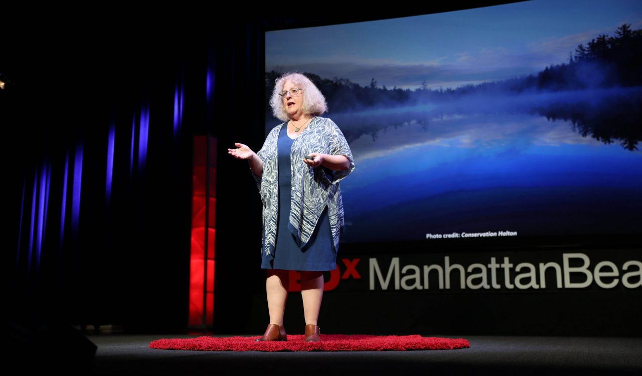 Long-distance view of Brock University Professor of Earth Sciences Francine McCarthy lit up, standing on a red carpet on a stage, with a large slide depicting Crawford Lake and lettering saying “TEDx ManhattanBeach.”