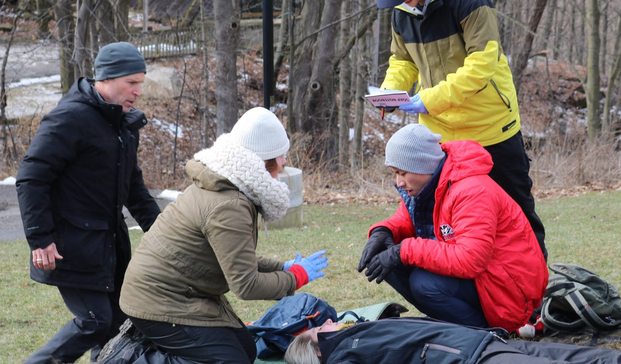 Outside in the wintertime, four people gather around a person laying on the floor simulating a medical distress situation.