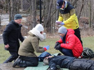 Outside in the wintertime, four people gather around a person laying on the floor simulating a medical distress situation.