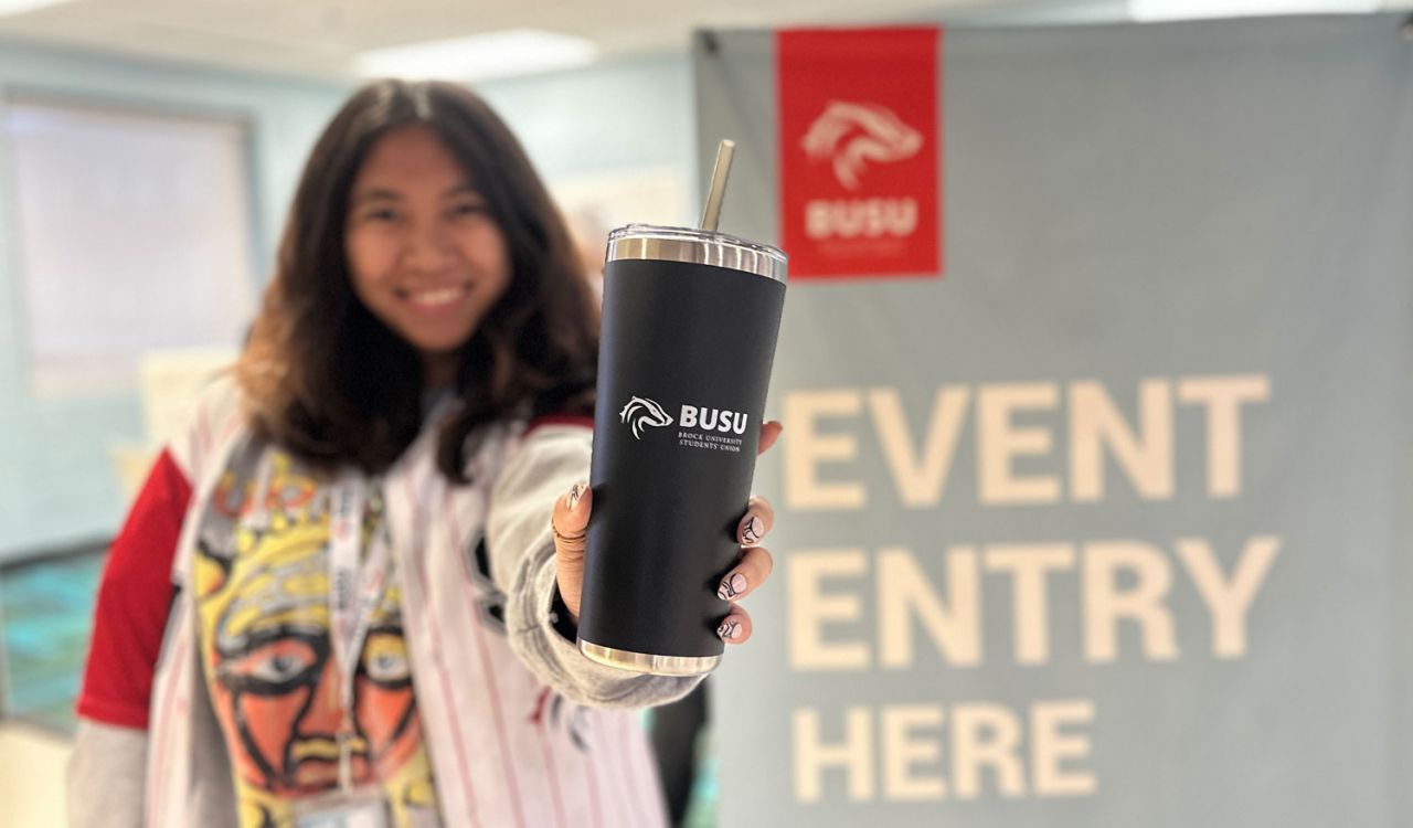 A person, who is out of focus, holds a black insulated tumbler with a Brock University Students’ Union logo printed on it in white.