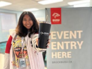 A person, who is out of focus, holds a black insulated tumbler with a Brock University Students’ Union logo printed on it in white.