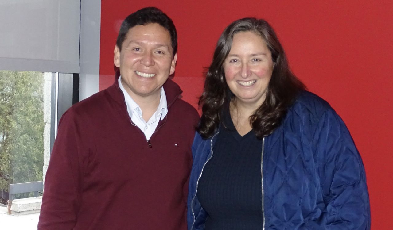 Two people pose for a photo against a red wall beside a window.