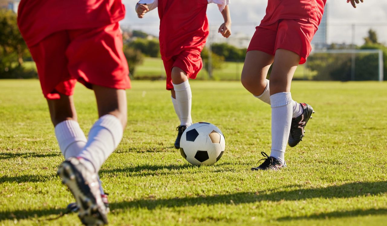 Three youths kick a soccer ball on an outdoor grass field. The image focuses on the lower half of their bodies, so all that is seen is the bottom of their red jersey tops, red shorts, white knee-high socks and black soccer cleats.