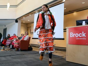 A First Nations woman in an orange jingle dress dances while a panel of women look on.