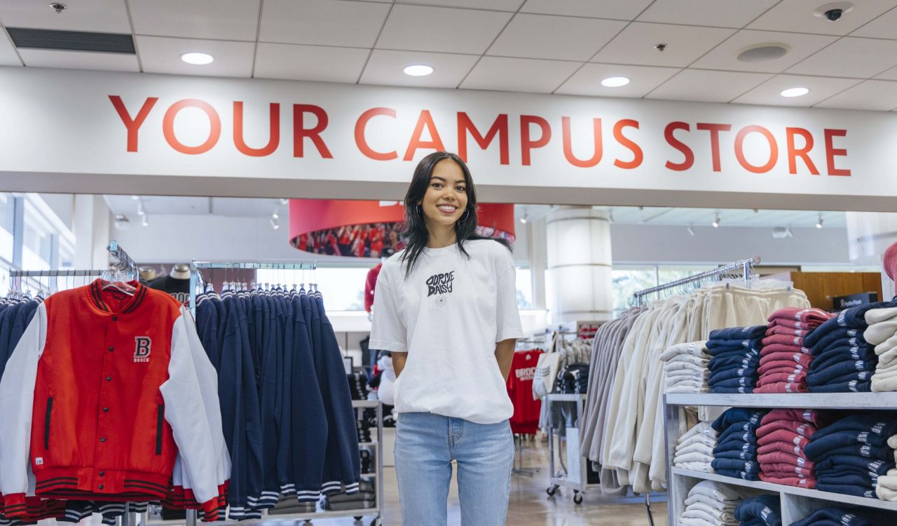 A young woman wearing a whit shirt and jeans stands in the Brock Campus Store surrounded by clothing displays.