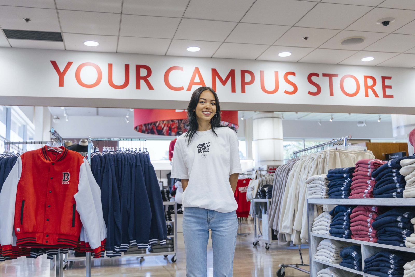 A young woman wearing a whit shirt and jeans stands in the Brock Campus Store surrounded by clothing displays.